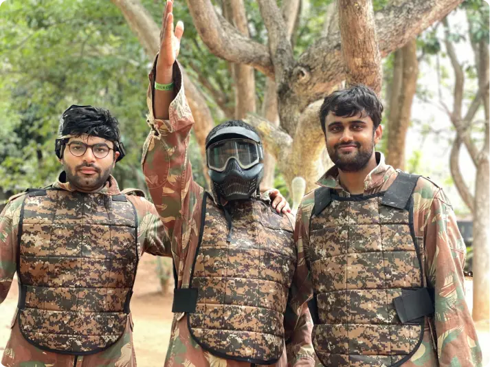 Three team members in camouflage gear for paintball, one raising hand victoriously, showcasing team-building activities