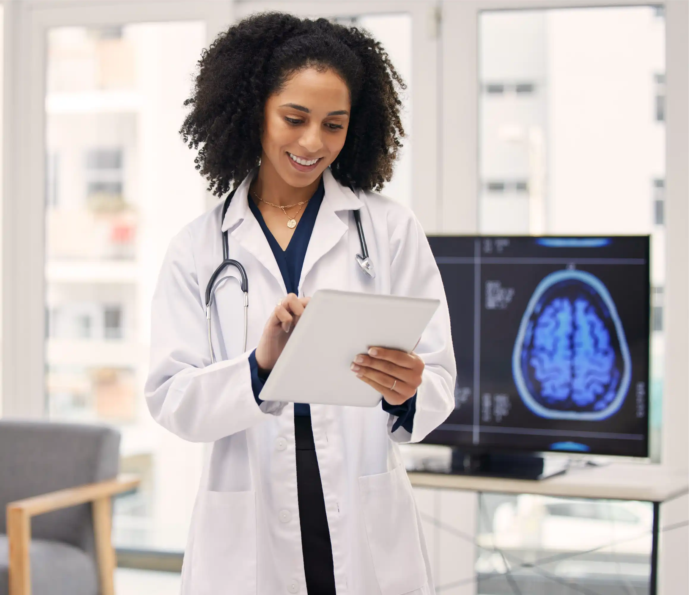 Smiling doctor with curly hair reviewing digital patient data on tablet, brain scan visible on monitor behind