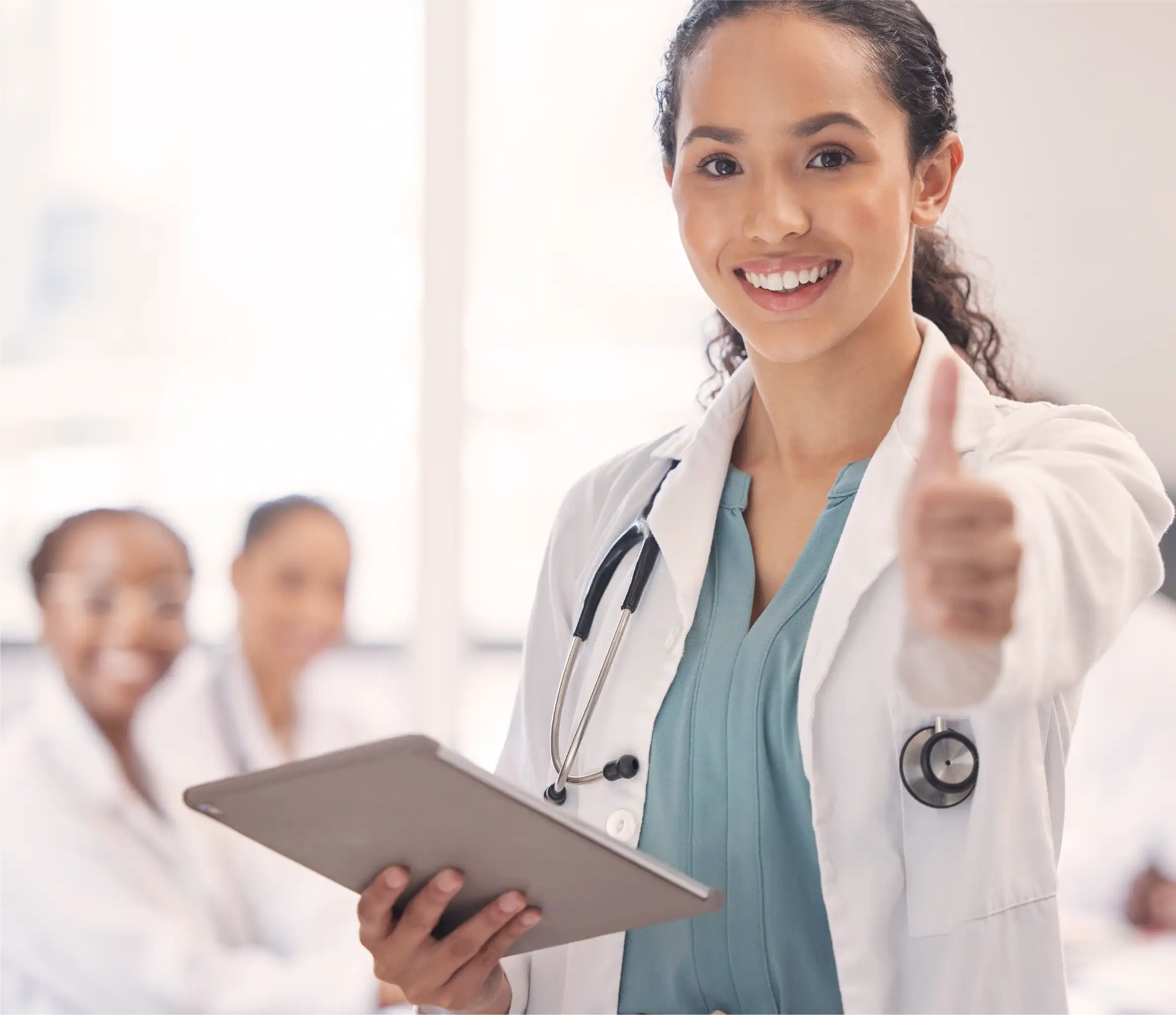Smiling female doctor with stethoscope holding tablet, giving thumbs up, with colleagues in background