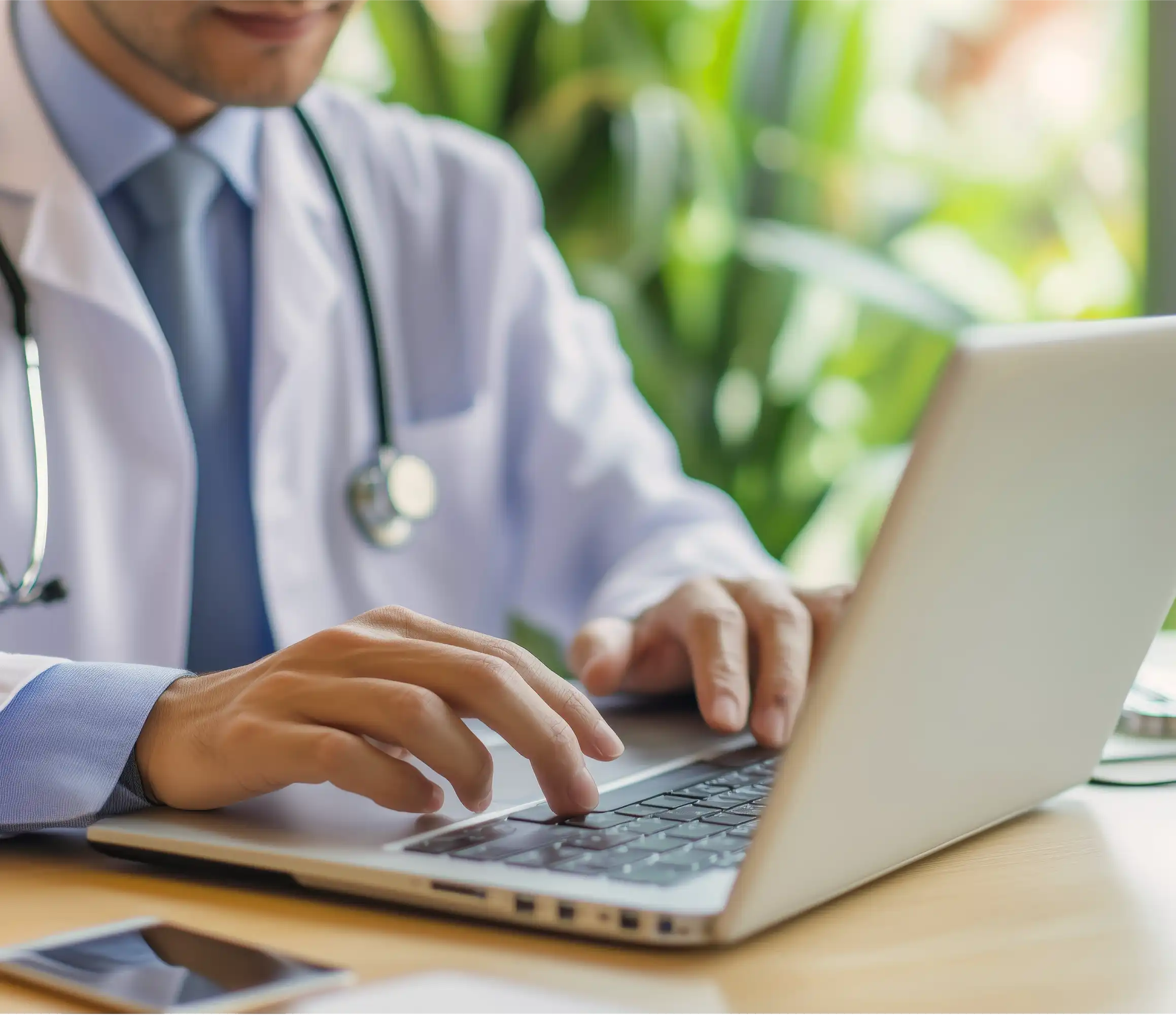 Doctor in white coat with stethoscope typing on laptop, green plants in background